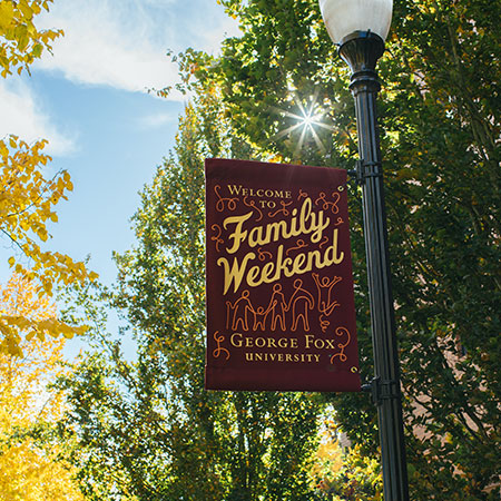 灯柱横幅上写着“欢迎来到乔治福克斯大学的家庭周末”，后面是树和阳光